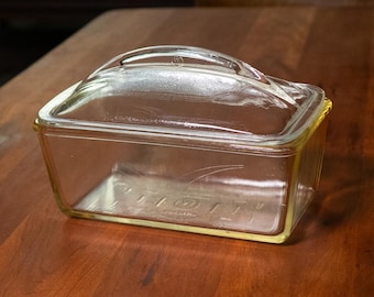 May include: A clear, rectangular glass baking dish with a handle on the lid, sitting on a dark brown wooden table. The dish has a yellow-tinted rim. The background shows a living room setting with furniture and decor.