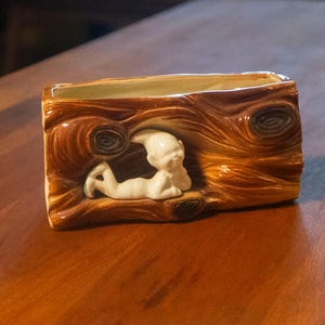 May include: A rectangular ceramic planter with a brown wood-grain texture. A white, reclining cherub figure is sculpted in the center. The planter is set on a polished wooden surface.