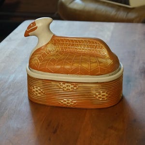 Puede incluir: Una caja decorativa de cerámica con tapa en forma de pájaro. La caja es principalmente naranja con detalles blancos y patrones intrincados. La tapa presenta la cabeza y el cuerpo de un pájaro, mientras que la base tiene un diseño texturizado.