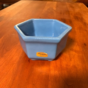 May include: A light blue, hexagonal ceramic bowl with a glossy finish. The bowl has a small, rectangular sticker with the word "Haeger" on it. The bowl is sitting on a wooden surface.