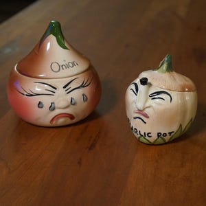 Può includere: Due contenitori in ceramica per cipolle e aglio con facce. Il vaso di cipolle è marrone, rosa e bianco, con la parola "Cipolla" in alto. Il vaso di aglio è bianco e verde, con le parole "Garlic Pot". Entrambi hanno facce tristi.