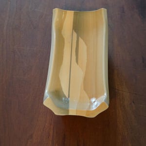 Könnte beinhalten: Eine rechteckige Glasschale mit gewölbter Ober- und Unterseite. Die Schale hat ein hellgelbes und beigefarbenes Farbschema mit einem vertikalen Design in der Mitte. Die Schale ist etwa 20 cm lang.