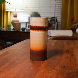 May include: A cylindrical ceramic vase with a gradient design. The base is dark brown, transitioning to orange, then light tan, with a dark brown band near the top. The vase sits on a wooden table. Home decor.