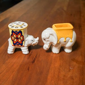 May include: Two white ceramic elephant figurines. One has a yellow and orange floral design on its back, and the other has a yellow container on its back. Both have a trunk and four legs.