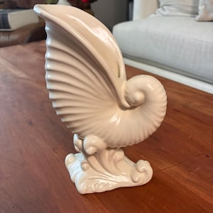 Puede incluir: Una escultura de cerámica de color crema con forma de concha de nautilus. La concha presenta un diseño detallado y acanalado y una forma en espiral. La base de la escultura tiene un diseño en forma de ola. La escultura se exhibe sobre una superficie de madera.