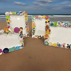 Könnte beinhalten: Vier weiße Bilderrahmen, verziert mit bunten Disney-Charakter-Pins. Die Rahmen sind rechteckig mit abgerundeten Ecken und stehen an einem Sandstrand mit dem Meer im Hintergrund.