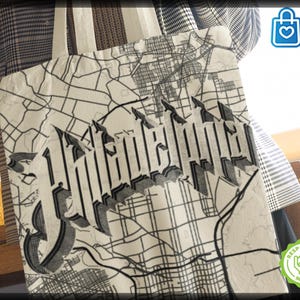 May include: Cream-colored tote bag featuring a black and white map design with the word "Wonderland" in a gothic font. The bag has a simple strap and is made of a light-colored fabric. The bag is being held by someone wearing a plaid blazer.