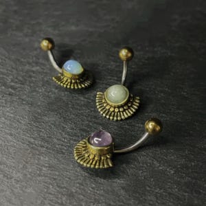 Pode incluir: Três piercings para o umbigo com uma barra curva prateada e uma bola dourada no topo. Cada anel tem uma pedra preciosa de cor diferente: azul, verde e roxo. A pedra preciosa é colocada numa base dourada em forma de leque.