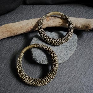 Op de afbeelding: Twee sierlijke, cirkelvormige oorbellen in bronskleur met een herhalend patroon. De oorbellen liggen op een grijze steen, met een stuk drijfhout op de achtergrond. De oorbellen hebben een vintage of tribale uitstraling, met een diameter van ongeveer 3 cm.
