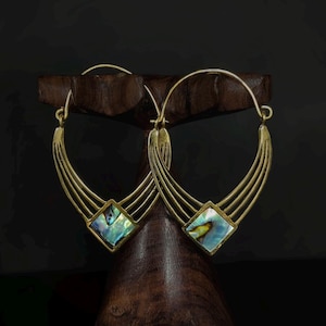 Op de afbeelding: Goudkleurige oorringen met een geometrisch ontwerp. Elke oorring heeft een vierkante abalone schelpinleg en meerdere gebogen lijnen. De oorbellen worden getoond op een donkerhouten standaard tegen een zwarte achtergrond.