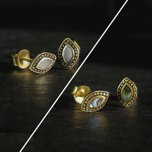 Può includere: Orecchini a bottone color oro con un design ovale a forma di occhio. Ogni orecchino presenta una pietra centrale iridescente, circondata da un bordo di perline. Gli orecchini sono esposti su una superficie scura.