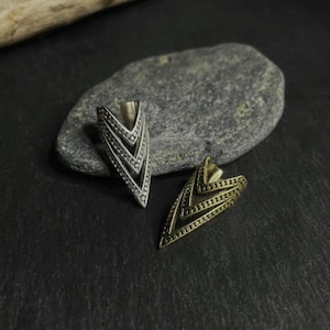 May include: Two metal arrow-shaped rings, one silver and one gold, are displayed on a dark surface. The rings have a layered design with intricate detailing. The silver ring is on the left, and the gold ring is on the right.