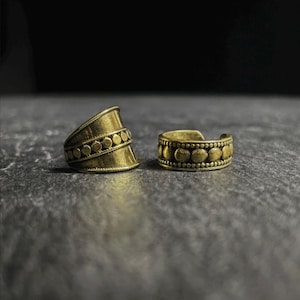 Pode incluir: Dois anéis dourados com um design texturizado. Os anéis apresentam uma série de pequenos círculos em relevo ao redor da banda. Um é uma banda larga, e o outro é um anel menor e ajustável. O fundo é escuro.