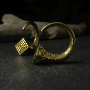 May include: Two brass-coloured earrings. One earring features a square with a spiral design, while the other has a triangular end with etched lines and a star pattern. The earrings have a curved, hook-like shape.