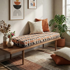 May include: A wooden bench with a patterned cushion in orange, black, and cream tones. Two throw pillows in cream and rust colours rest on the bench. A rug with a geometric pattern and a potted plant are also in the image. Decorative vases and framed art complete the room.