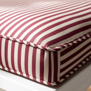 Può includere: Primo piano di un cuscino con un motivo a righe bordeaux e bianche. Il cuscino ha un design spesso e imbottito con un bordo su misura. Le strisce sono verticali e uniformemente distanziate, creando un'estetica classica e nautica. Il cuscino è probabilmente per mobili da esterno.