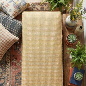 May include: A rectangular yellow bench with a grid pattern is surrounded by decorative pillows, plants, and a wooden table. The pillows have various patterns and colors. The table holds potted plants, books, and vases with flowers.
