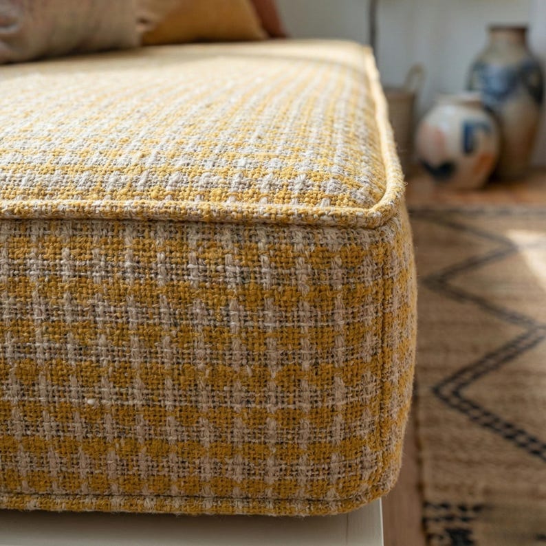 May include: Close-up of a yellow and beige upholstered ottoman. The fabric has a woven texture with a repeating pattern. The edges are neatly finished with a matching trim. The ottoman is part of a home decor collection.