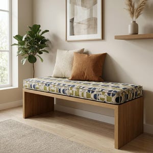 May include: A wooden bench with a patterned cushion in shades of blue, green, and beige. Two throw pillows in cream and rust-orange rest on the bench. The bench is in a room with a plant and artwork.