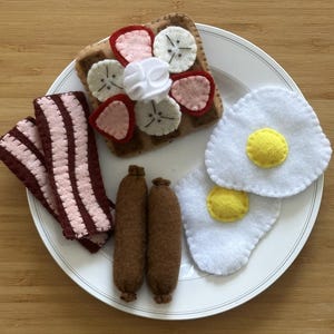 Puede incluir: Un juego de desayuno de fieltro sobre un plato blanco. El juego incluye un gofre cubierto con fruta de fieltro, dos huevos fritos, dos salchichas y tiras de tocino. La comida de fieltro está dispuesta en un plato blanco.