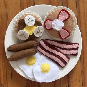 May include: A felt breakfast set on a white plate. The set includes pancakes with bacon and whipped cream, a pancake with banana slices, two sausages, two fried eggs, and two strips of bacon. The felt items are in shades of brown, white, yellow, and pink.