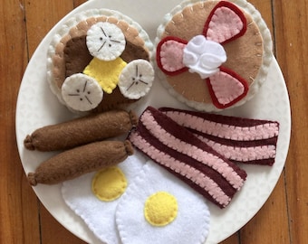 May include: A felt breakfast set on a white plate. The set includes pancakes with bacon and whipped cream, a pancake with banana slices, two sausages, two fried eggs, and two strips of bacon. The felt items are in shades of brown, white, yellow, and pink.