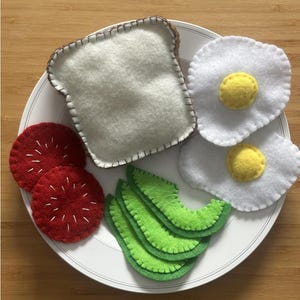 Może przedstawiać: Zestaw śniadaniowy z filcu na białym talerzu. Zestaw zawiera kromkę tostową, dwa smażone jajka, dwa plasterki pomidora i plasterki awokado. Tost jest jasnobrązowy z białymi przeszyciami. Jajka mają żółte żółtka. Pomidory są czerwone z białymi przeszyciami.