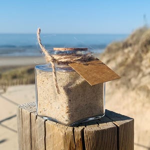 以下が含まれることがあります： 砂で満たされたガラス瓶が、紐と茶色の紙タグで結ばれています。瓶は木製の支柱の上に置かれ、背景にはビーチと青い空がぼやけています。