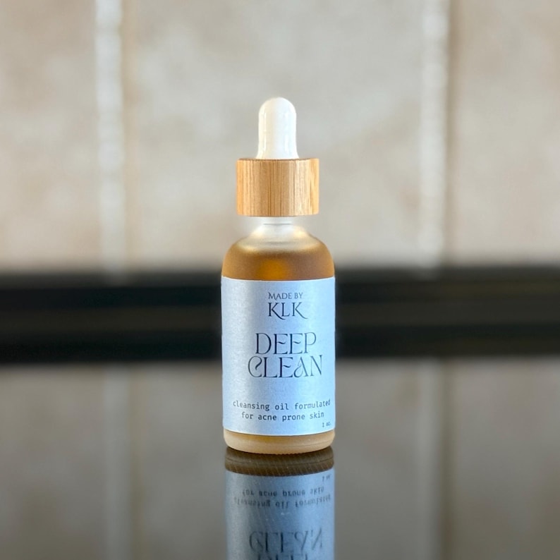 May include: A glass bottle with a wooden cap and a light blue label that reads "MADE BY KLK DEEP CLEAN cleansing oil formulated for acne prone skin 1 fl oz".