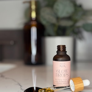 Puede incluir: Una botella de vidrio marr&oacute;n de aceite facial antiedad "GLOW DROPS" con un cuentagotas y una peque&ntilde;a cuchara plateada. La etiqueta dice "MADE BY KLK" y enumera los ingredientes. El cuentagotas es de bamb&uacute; y vidrio. La cuchara contiene un poco de aceite.