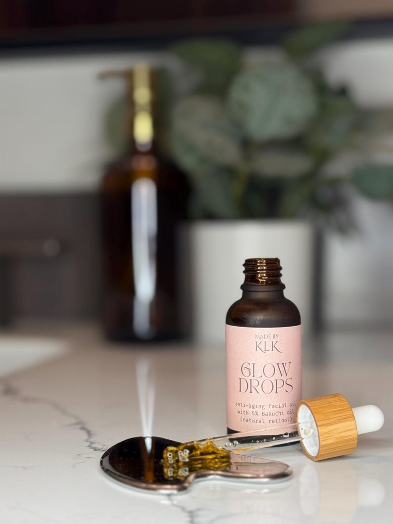 Puede incluir: Una botella de vidrio marr&oacute;n de aceite facial antiedad "GLOW DROPS" con un cuentagotas y una peque&ntilde;a cuchara plateada. La etiqueta dice "MADE BY KLK" y enumera los ingredientes. El cuentagotas es de bamb&uacute; y vidrio. La cuchara contiene un poco de aceite.