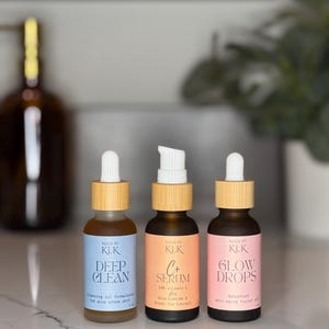 3 bottles in skincare line. The cleansing oil is a frosted glass bottle with a blue label and white and bamboo dropper. The middle bottle, vitamin c serum, has amber glass with an orange label and bamboo and white pump top. The third bottle, Glow Drops, has amber glass bottle with a pink label and bamboo and white dropper top. They are sitting on a bathroom counter with a plant and soap pump in the background.