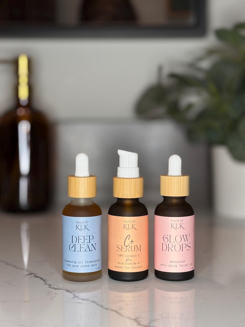 3 bottles in skincare line. The cleansing oil is a frosted glass bottle with a blue label and white and bamboo dropper. The middle bottle, vitamin c serum, has amber glass with an orange label and bamboo and white pump top. The third bottle, Glow Drops, has amber glass bottle with a pink label and bamboo and white dropper top. They are sitting on a bathroom counter with a plant and soap pump in the background.
