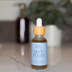 Puede incluir: Una peque&ntilde;a botella de vidrio transparente con tapa de madera y cuentagotas blanco. La etiqueta dice "DEEP CLEAN" y "cleansing oil formulated for acne prone skin". La botella contiene un l&iacute;quido dorado. La marca es "KLK".