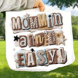 Puede incluir: Un bolso tote blanco con el texto "MOMMIN AIN'T EASY!" en un diseño de patchwork. Las letras son en tonos marrón, beige y azul, con detalles de estrellas. El bolso se sostiene contra un fondo verde borroso.