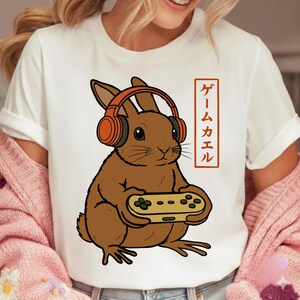 Könnte beinhalten: Weißes T-Shirt mit einem braunen Hasen im Cartoon-Stil, der orangefarbene Kopfhörer trägt und einen Videospiel-Controller hält. Japanischer Text ist auf der rechten Seite des Hasen aufgedruckt.
