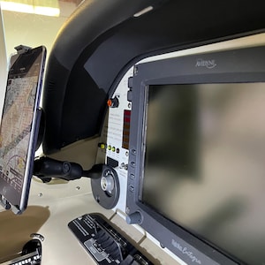 May include: A close-up of a cockpit interior with a tablet mounted on the left side and a large screen on the right. The screen displays the text "Avidyne" and "Pak Entegra". The screen is surrounded by a black bezel and has a gray background. There are several buttons and switches below the screen.