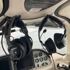 May include: Two black headsets with microphones are hanging from the ceiling of a small aircraft. The headset on the left has the word "BOSE" printed on the side. The headsets are likely used for communication during flight.