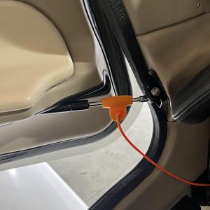 May include: An orange T-shaped tool with a red cord is inserted into a car door mechanism. The car door interior is beige and black, with a white door panel visible in the background. The tool appears to be a safety device or a repair component.