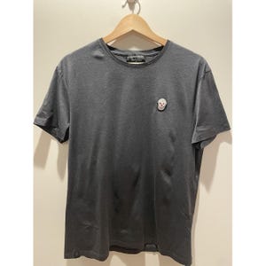 May include: A dark grey t-shirt featuring a small skull motif on the chest. The top has short sleeves and a crew neck. The brand name is printed on the collar.