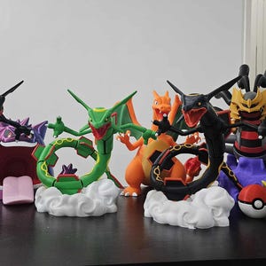 May include: A collection of colorful Pokémon figurines, including Rayquaza, Charizard, and Meowth, displayed on a dark surface. The figures feature various colors and designs, with some posed on cloud-shaped bases. Other items include Poké Balls and a Team Rocket emblem.
