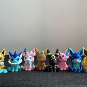 Può includere: Una collezione di otto figurine colorate di Pokémon. Le figure includono personaggi in tonalità di giallo, blu, rosa, marrone e nero. Ogni figura ha caratteristiche uniche, tra cui orecchie, code ed espressioni facciali. Sono disposte in fila.