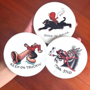 May include: Set of three white ceramic coasters with vintage tattoo-style illustrations. Designs include a devil riding a panther, a man walking with the text "Keep on Truckin'," and a horse smoking a cigarette with the text "Cool Stud."