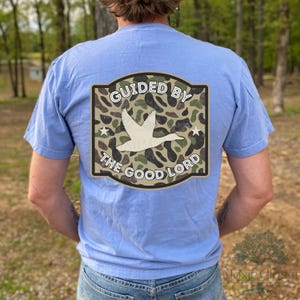 Könnte beinhalten: Hellblaues T-Shirt mit Grafik auf dem Rücken. Die Grafik zeigt eine weiße Ente im Flug über einem Tarnmuster, mit den Worten "GUIDED BY THE GOOD LORD" in einem geschwungenen Design.