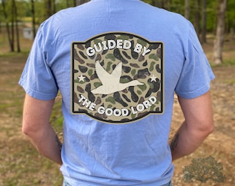 May include: A light blue t-shirt with a graphic on the back. The graphic features a white duck in flight over a camouflage pattern, with the words "GUIDED BY THE GOOD LORD" in a curved design.
