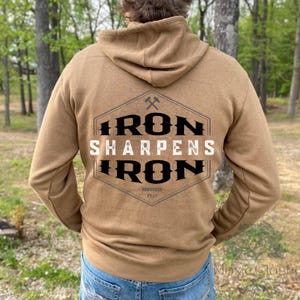 May include: A tan hoodie with a hood and long sleeves. The back of the hoodie features a graphic with the text "IRON SHARPENS IRON" in black and white, with a small axe symbol above the text. The text "PROVERBS 27:17" is below.