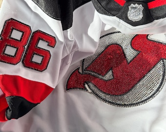May include: White hockey jersey with black and red accents. Features the number "86" in red on the sleeve and a red and silver logo on the front. The jersey has a black collar with red trim and a silver NHL logo.