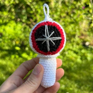 May include: A white crocheted ornament with a circular design. It has a black centre, a red border, and a grey starburst. The ornament has a white handle and a hanging loop. The background is blurred green foliage.