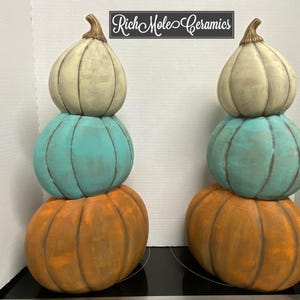 May include: Two stacked ceramic pumpkins in shades of orange, teal, and cream. The pumpkins are arranged in a vertical tower, with the largest at the base and the smallest at the top. The pumpkins have a rustic, textured finish.