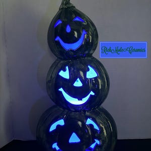 May include: A vertical stack of three dark, marbled ceramic pumpkins with cut-out jack-o'-lantern faces illuminated from within by a bright blue light. The text Rich Mole Ceramics is visible in a blue box to the right of the middle pumpkin.